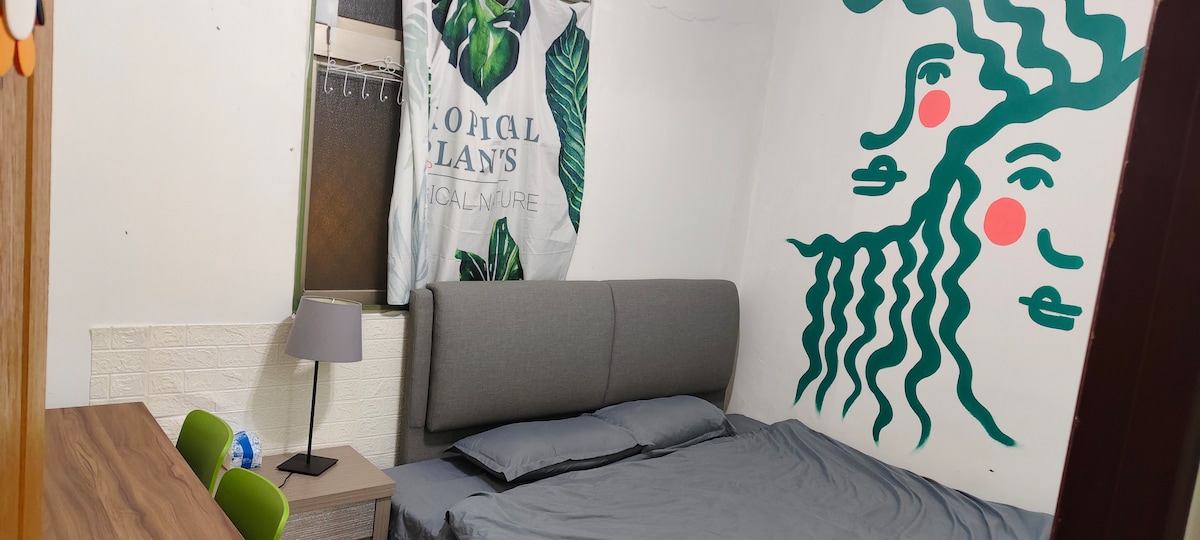 A bedroom features a spacious bed with a gray headboard and a soft bedding set. A wooden desk with two green chairs is situated nearby. A lively green mural adorns the wall, while natural light filters through a window dressed with a leafy curtain.