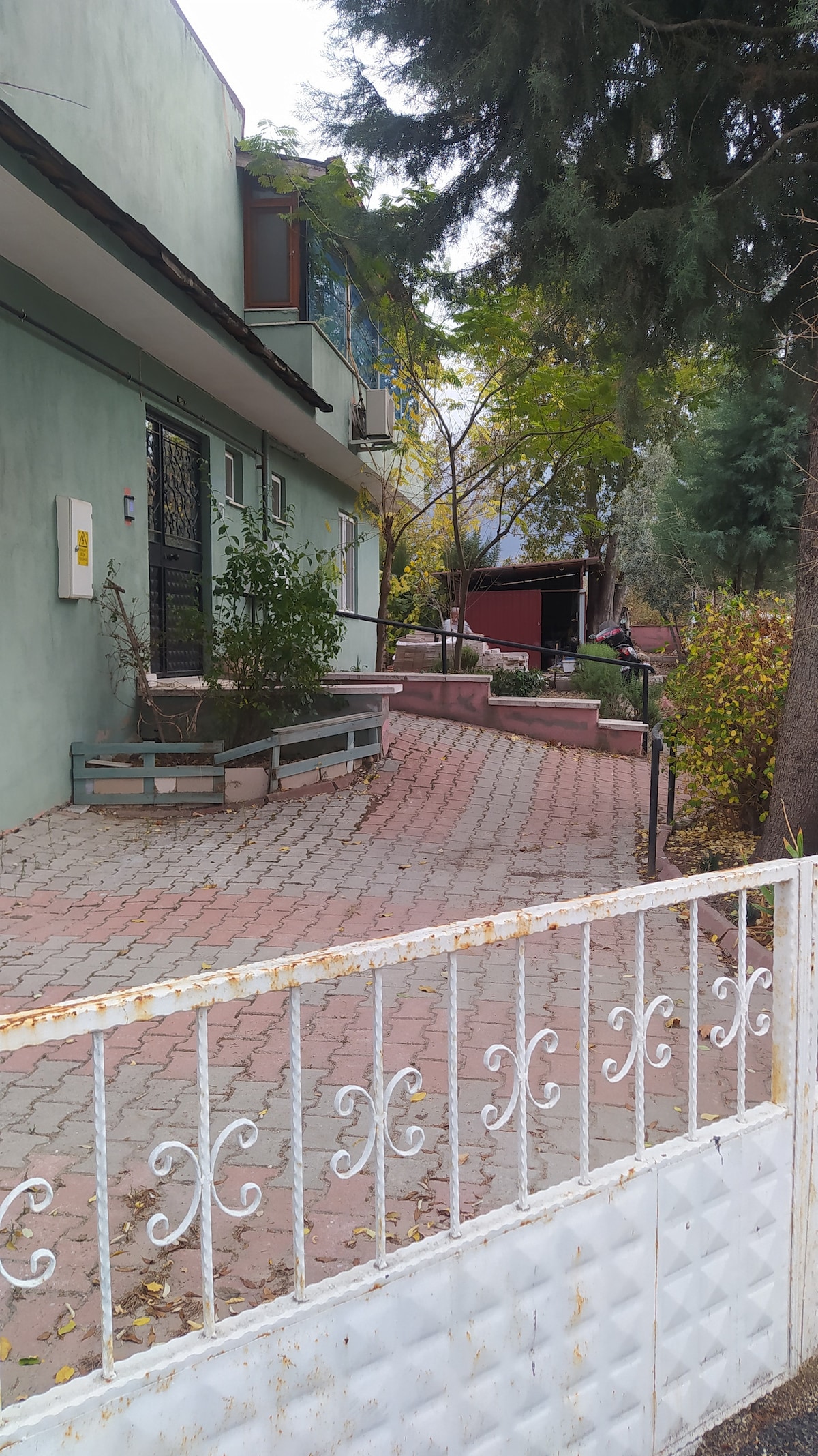 A pathway lined with potted plants leads to a two-story building, showcasing a green exterior wall. The entrance features stairs leading up, and a white metal fence borders the property. Surrounding trees provide shade in this welcoming outdoor space.