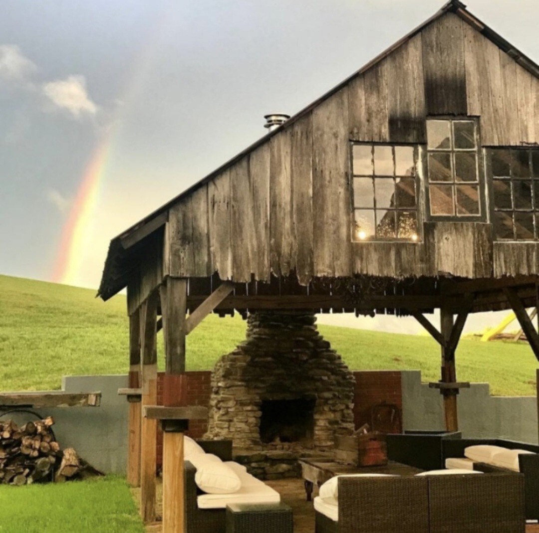 The rustic cabin features a wooden structure with a stone fireplace beneath a spacious overhang. A rainbow arches in the sky above, while a grassy hillside is visible in the background. Outdoor seating is arranged around the fireplace, complementing the natural surroundings.