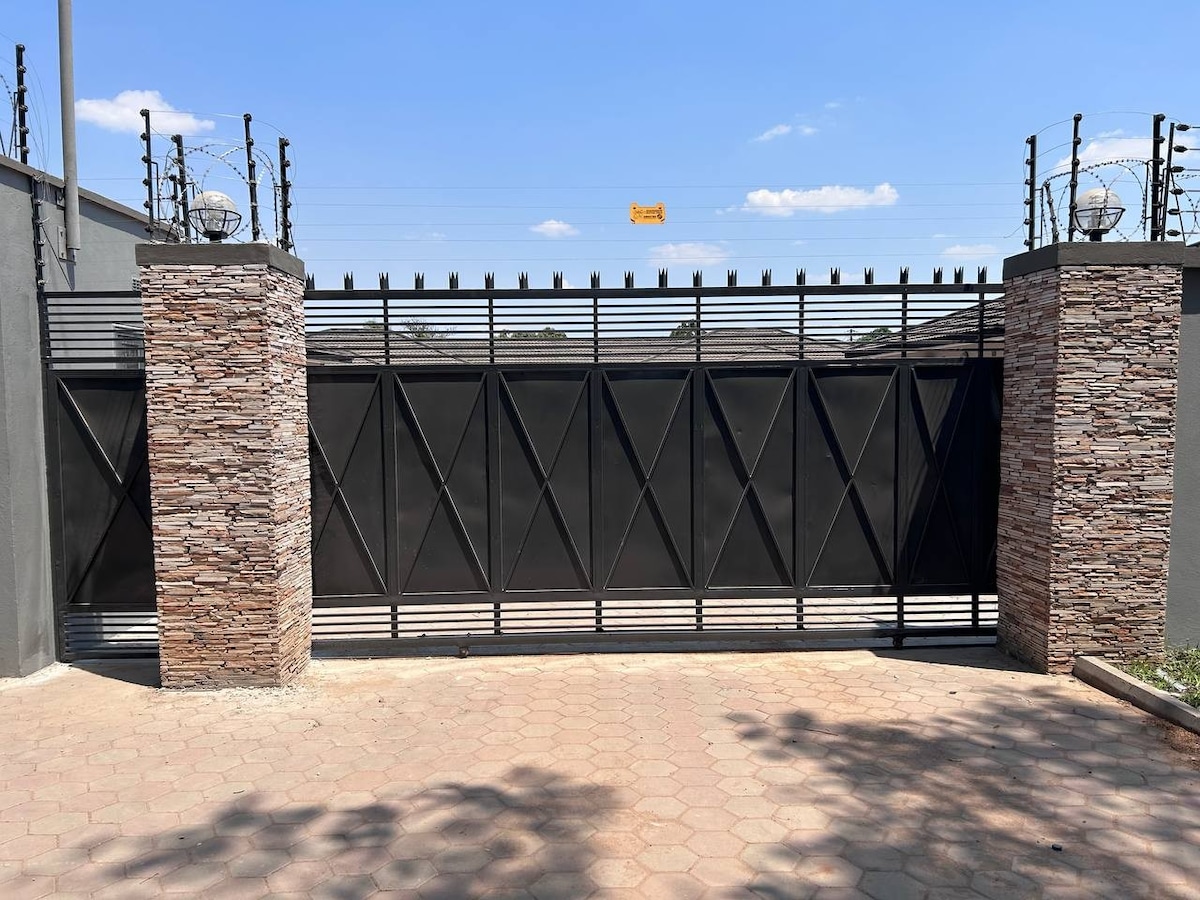 A secure entrance is presented by a tall black gate framed by stone pillars. The gate features a geometric design and is flanked by decorative lighting fixtures. A clear blue sky serves as the backdrop, with a paved pathway leading up to the gate.
