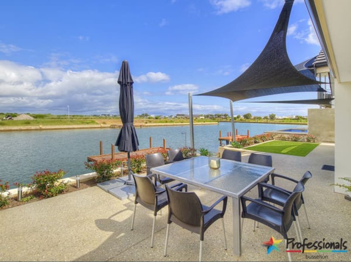 A patio area features a metal dining table surrounded by four dark chairs. A large umbrella provides shade, complemented by a sail shade overhead. The serene water view showcases a jetty and green space in the background, with flowers lining the patio edge.