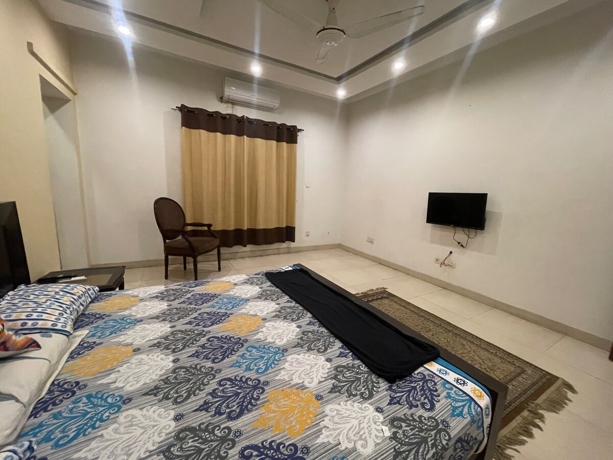 A spacious bedroom features a large bed with a colorful quilt draped across it. A sleek TV is mounted on the wall opposite. A single wooden chair sits in one corner, with natural light filtering in through large curtains. The floor is tiled, offering a clean and modern look.