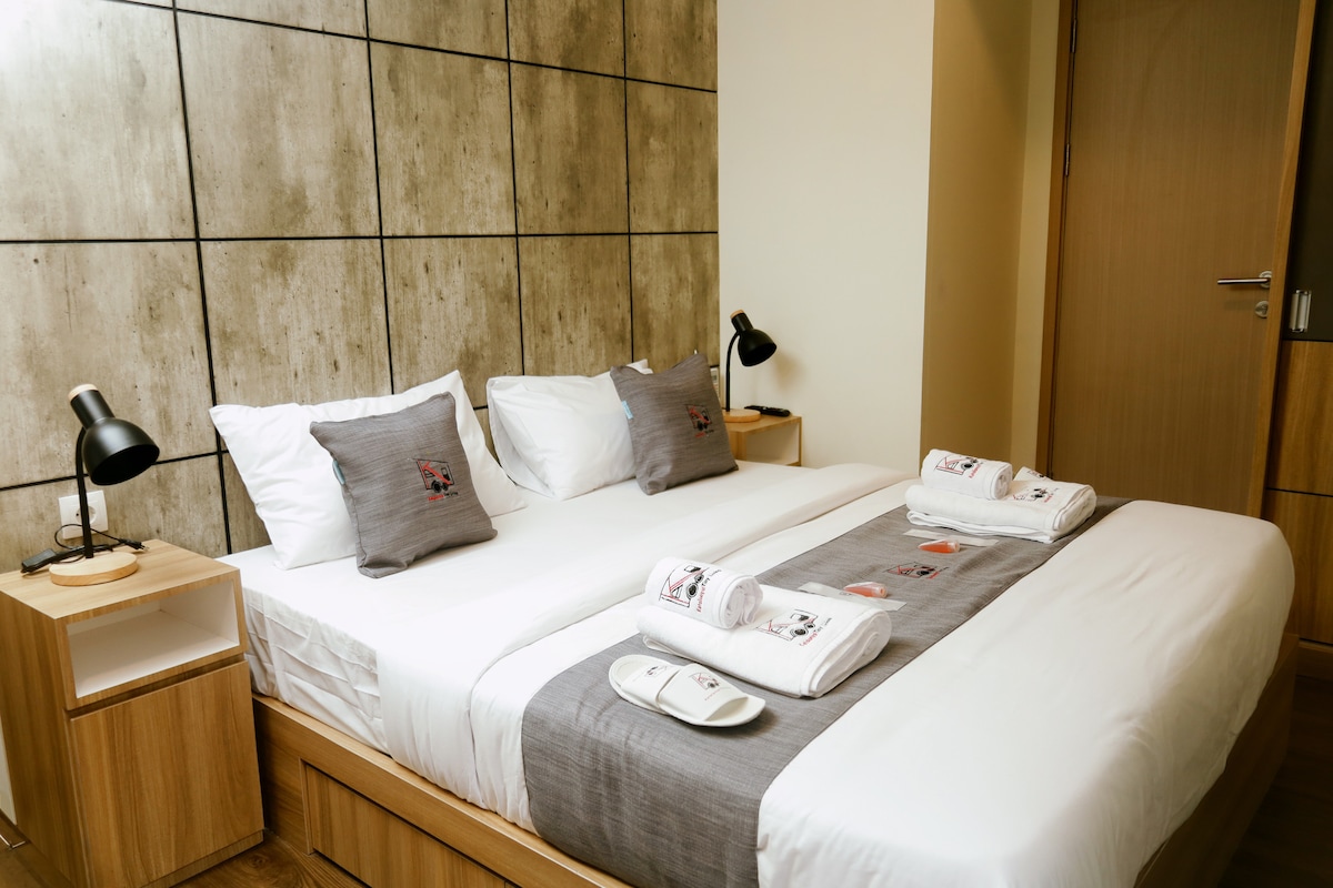 A well-appointed bedroom features a spacious bed with light linens, complemented by decorative pillows. A gray runner with neatly arranged towels adds a touch of detail. Two bedside lamps provide soft lighting against a textured wall, contributing to a serene environment.