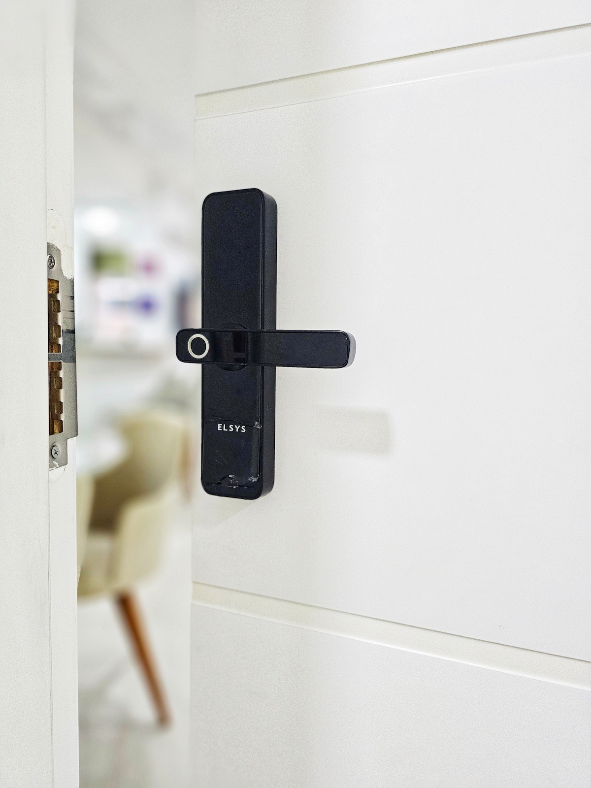 A modern door lock is featured prominently on a white door. The sleek black design incorporates a round keyhole and a simple lever handle, emphasizing security and ease of access. The interior glimpse shows soft colors, enhancing a sense of openness.