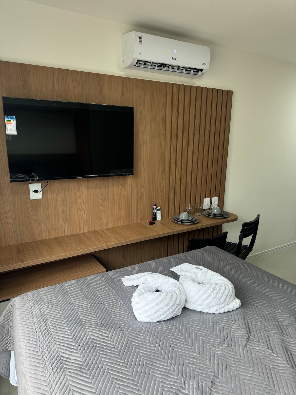 A neatly arranged room features a spacious bed dressed with a textured gray blanket. Two towel swans rest on the bed. A wooden wall unit holds a flat-screen television, while a small table is set for two, complete with glasses and a black chair.