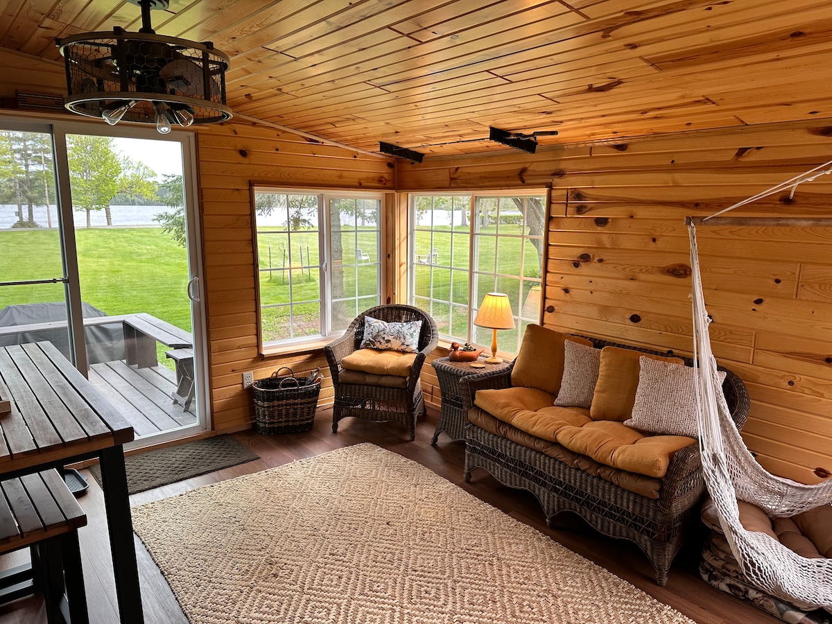 A sunlit interior space features wooden walls and ceiling, complemented by a cozy seating area with a wicker couch and a hammock. A table stands nearby, while large windows provide views of the outside greenery and lake, enhancing the inviting atmosphere.
