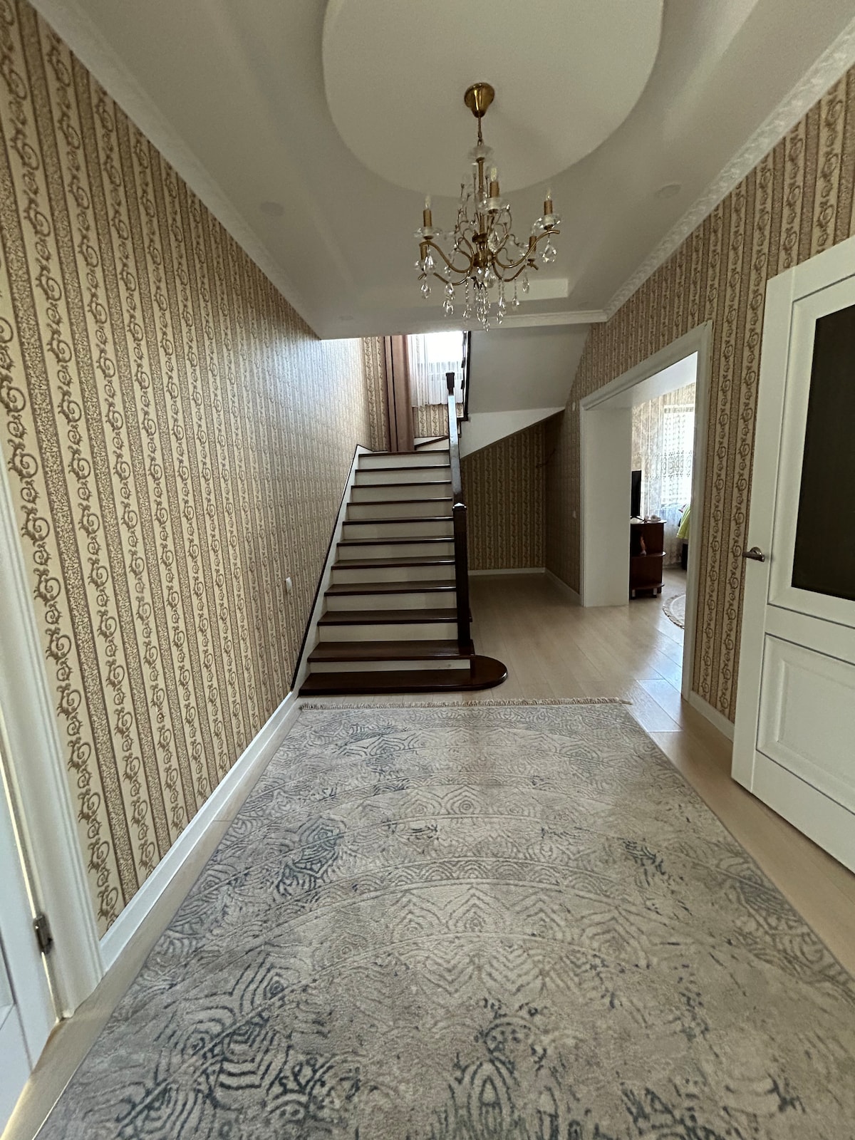 A well-lit hallway features patterned wallpaper and a soft area rug. A decorative chandelier hangs from the ceiling, illuminating the space. A staircase leads to the upper level, while a white door is visible on the right, enhancing the overall layout.