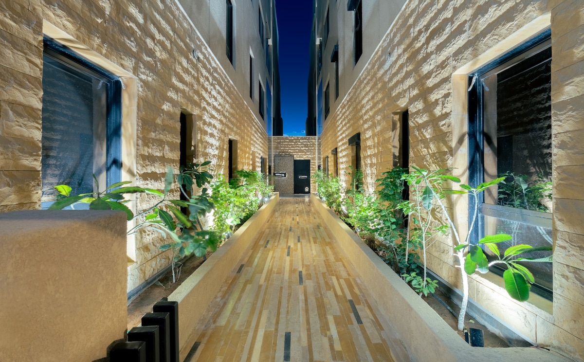 A narrow outdoor corridor is lined with textured stone walls and illuminated by soft lighting. Plants are positioned along the edges, providing greenery against the natural stone. The floor features a wooden surface with a varied pattern, leading towards a doorway at the far end.