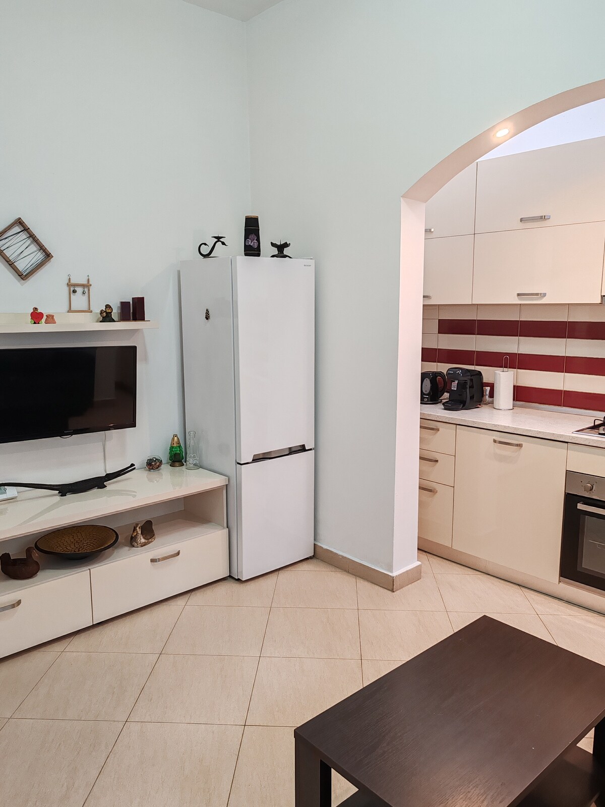 An open space kitchen and living area is shown, featuring a white refrigerator and a compact TV mounted on a light-colored unit. Red and white stripes decorate the kitchen walls, while a dark wooden table is positioned in front of the seating area.