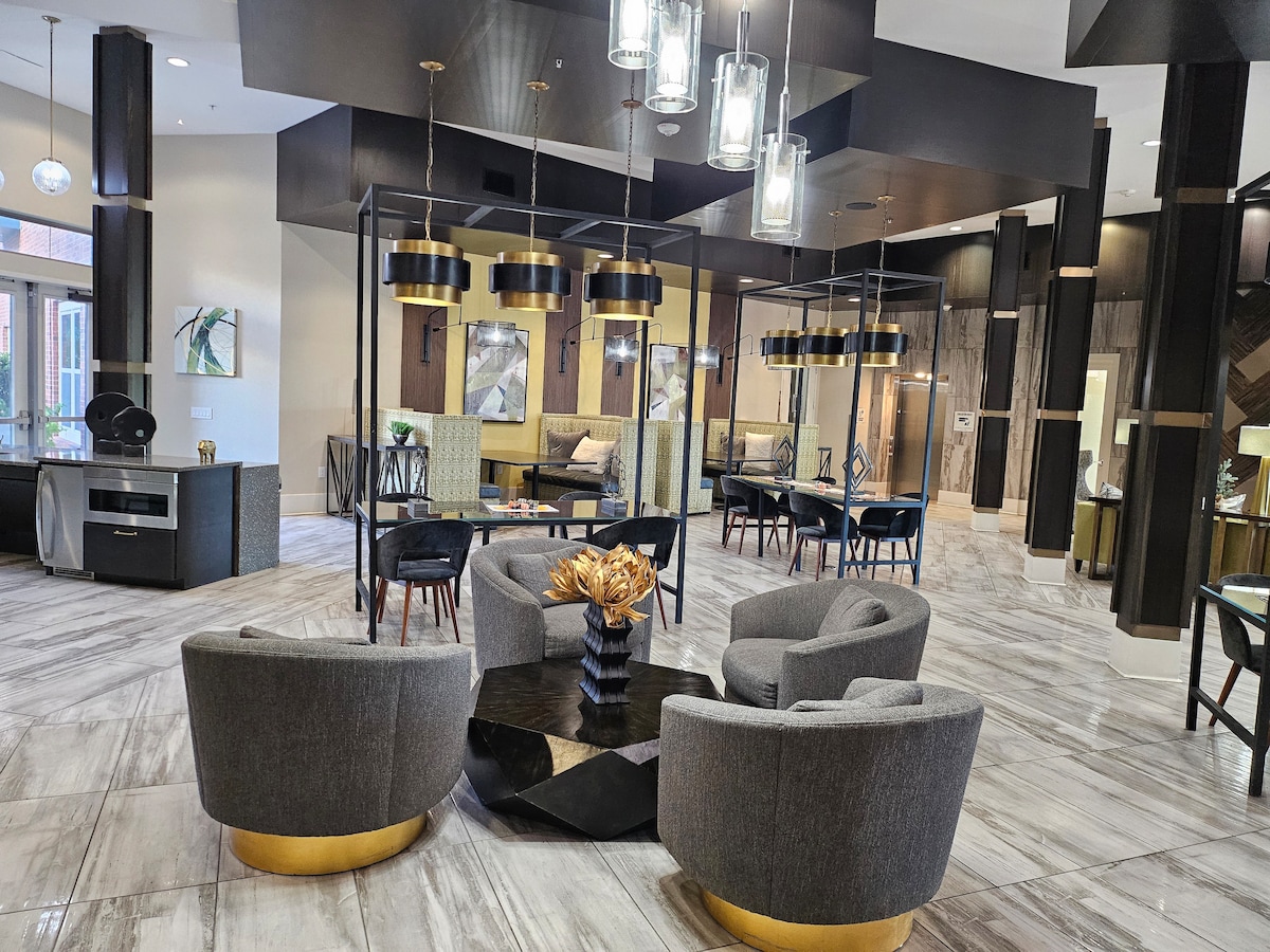 An open lobby area features contemporary seating arrangements, including two plush gray chairs and a black coffee table with a decorative centerpiece. Pendant lights illuminate the space, while a combination of textures and colors creates an inviting environment for relaxation or conversation.