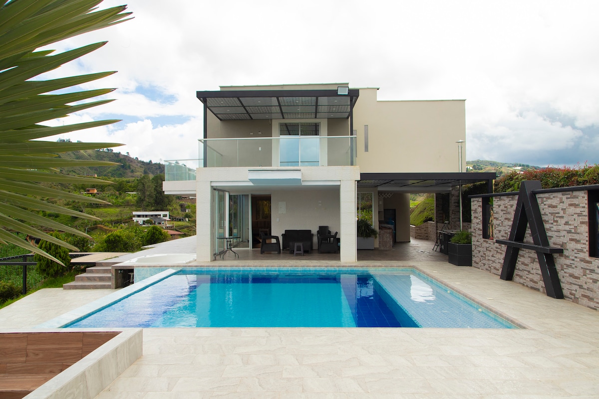 The modern exterior of the house is highlighted by a spacious swimming pool, surrounded by a sun deck. Large glass doors invite natural light to the interior spaces, with a covered patio area featuring outdoor seating. Lush greenery frames the serene landscape.