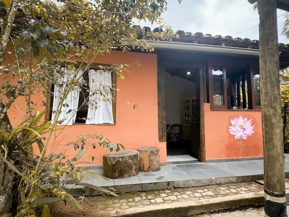 The exterior of the cottage features warm orange walls and a stone pathway. A small porch area is highlighted by wooden stools. Large windows allow natural light into the space, while lush greenery frames the entrance, enhancing the serene environment.