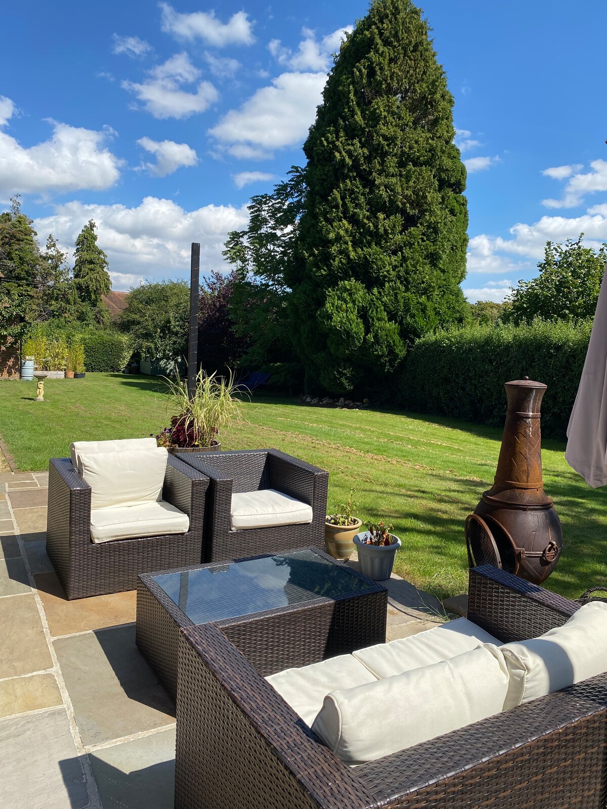 An outdoor patio area is furnished with two wicker chairs and a glass-topped table, providing a comfortable space for relaxation. A tall tree and greenery create a backdrop, with a chiminea positioned nearby. Bright skies and fluffy clouds enhance the serene atmosphere.