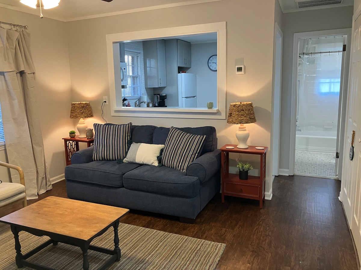 A cozy living area features a navy blue sofa with decorative pillows, flanked by two side tables with lamps. A wooden coffee table sits in front of a textured rug. Natural light is filtered through window treatments, creating a warm atmosphere. The bathroom door is visible in the background.