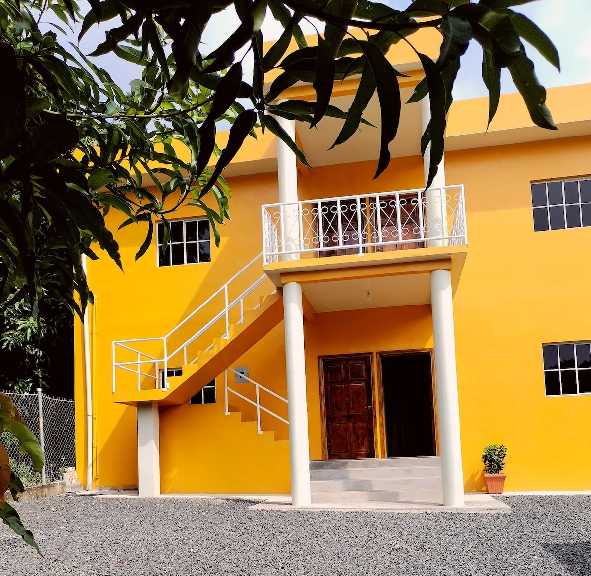 A bright yellow exterior features a two-story building with a welcoming entrance and a staircase leading to the upper level. Large windows provide natural light, and a potted plant is positioned near the door, enhancing the entryway.
