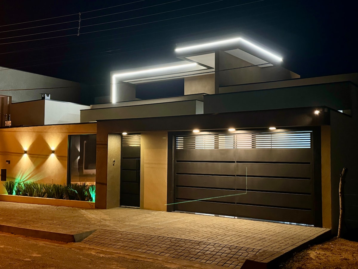 The modern exterior of the house features sleek lines and a contemporary design, illuminated by subtle outdoor lighting. A spacious driveway leads to an automatic garage door, while strategically placed greenery enhances the aesthetic at night.