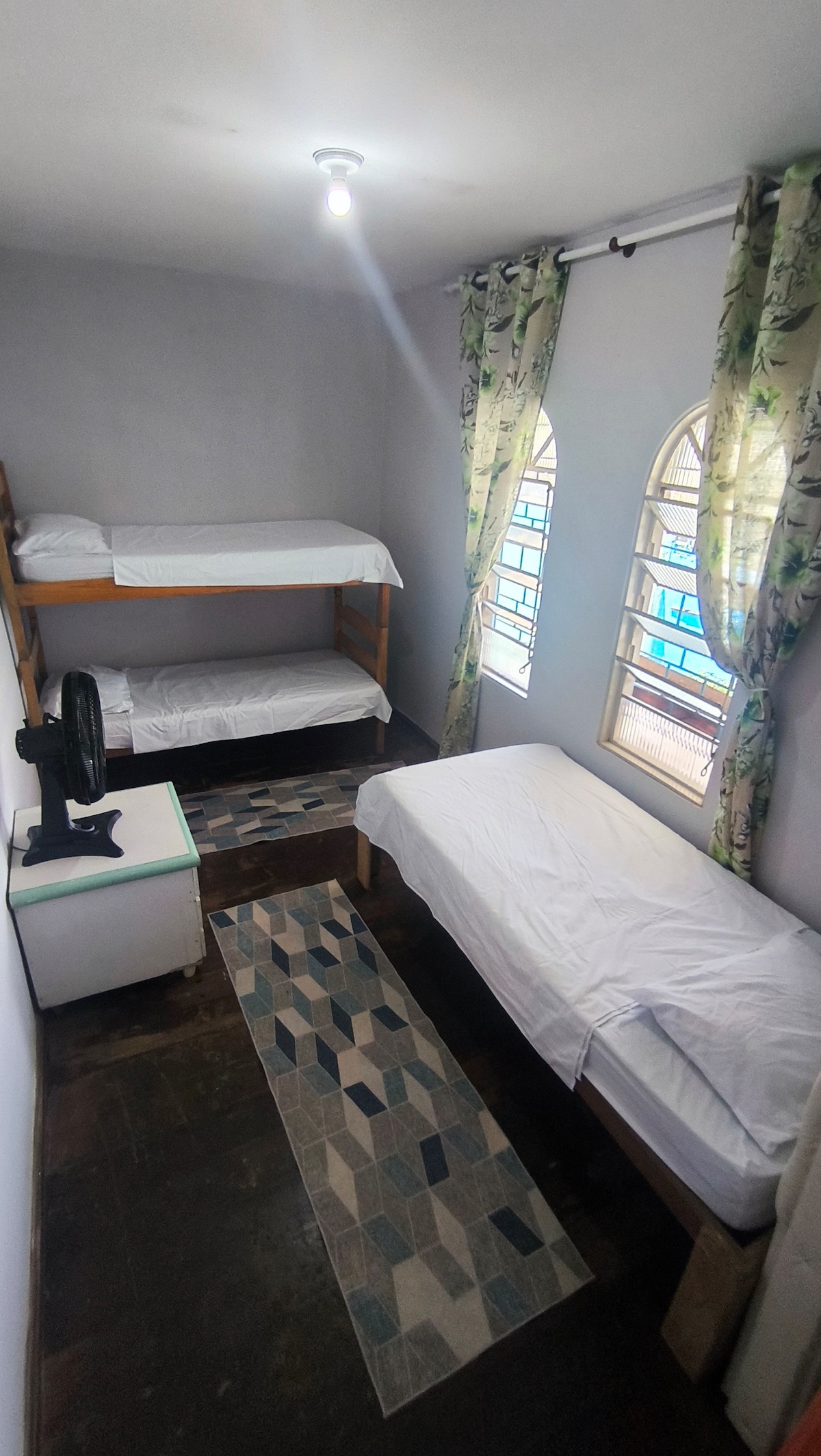 A shared room features a bunk bed and an additional single bed, arranged to maximize floor space. Two arched windows allow natural light to fill the room, complemented by light floral curtains. A small fan is positioned on a bedside table, and a patterned rug adds visual interest.