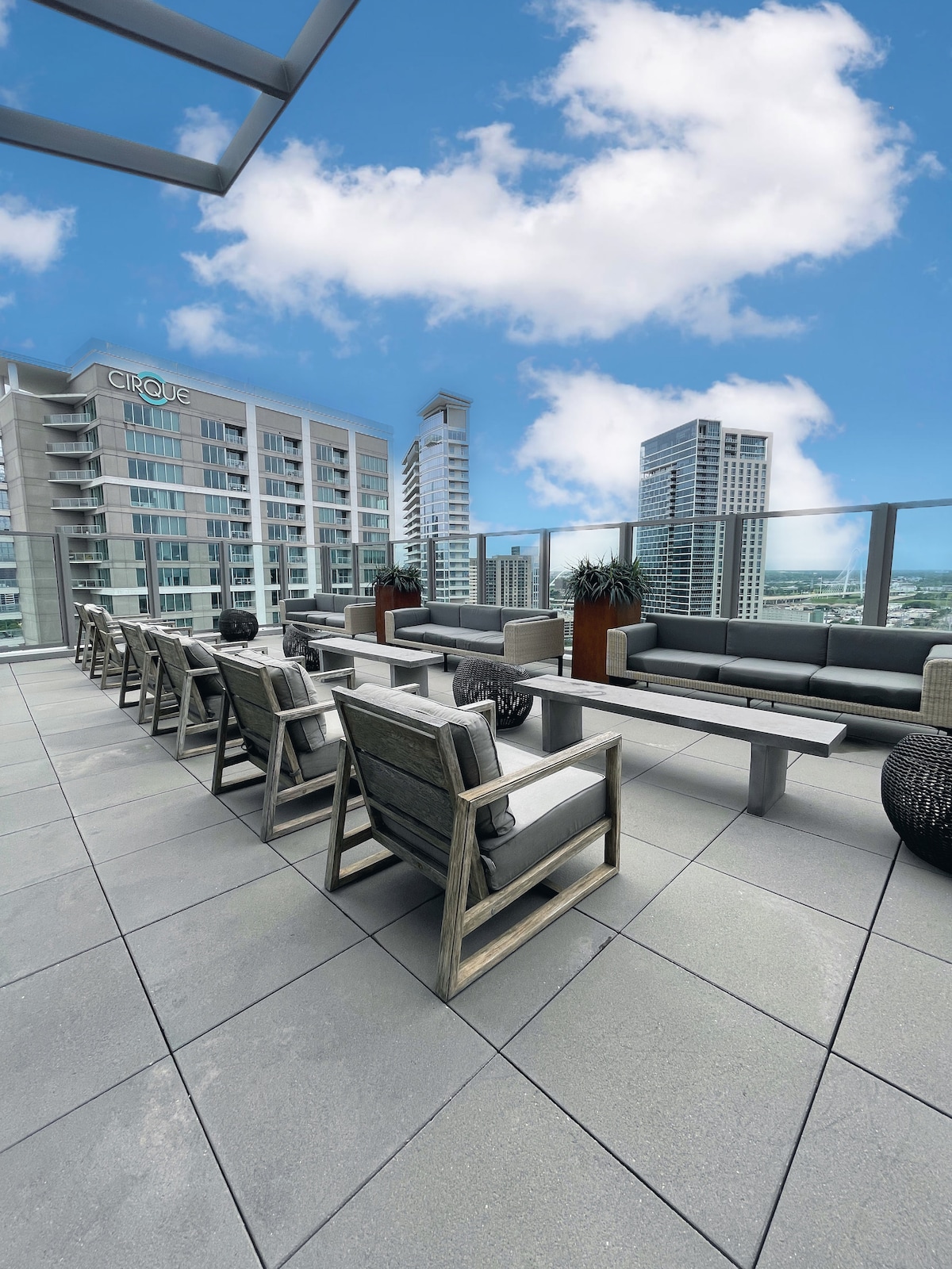 An outdoor lounge area is featured on the rooftop, furnished with multiple wooden chairs and long tables. Planters with greenery are placed throughout, and expansive city views can be seen in the background against a bright blue sky.