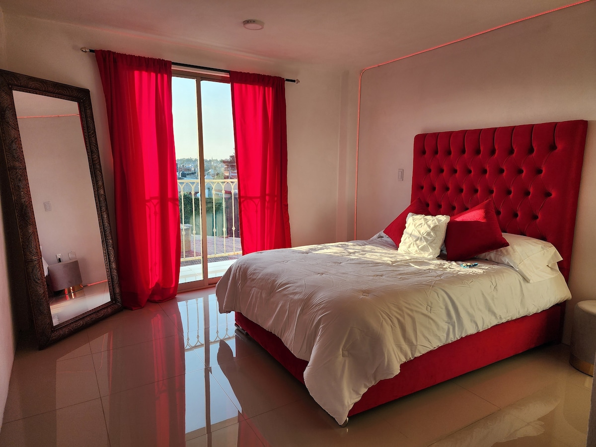 A spacious bedroom features a vibrant red velvet headboard framing a large bed adorned with pillows. Bright natural light floods the room through a sliding glass door, which leads to a balcony. A full-length mirror complements the decor, reflecting the room's elegant design.
