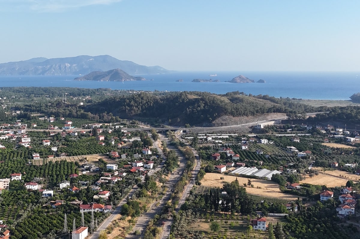 A panoramic view captures rolling hills and the sea in the distance. Lush green orchards stretch across the landscape, dotted with residential homes. The coastline features several islands visible on the horizon, while a clear blue sky enhances the serene atmosphere.
