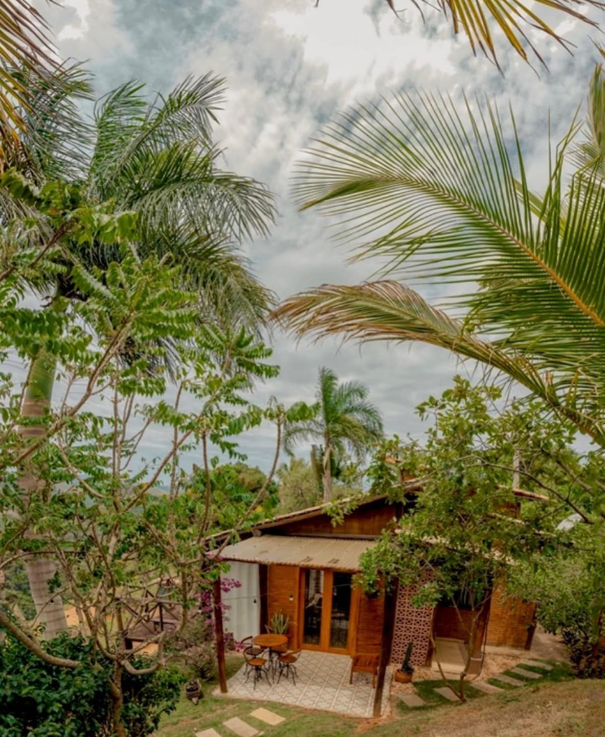 A charming wooden house is surrounded by lush greenery and tall palm trees. An outdoor seating area with a small table and chairs is visible on the patio, providing a serene view of the landscape. The structured pathways lead through the vibrant garden.