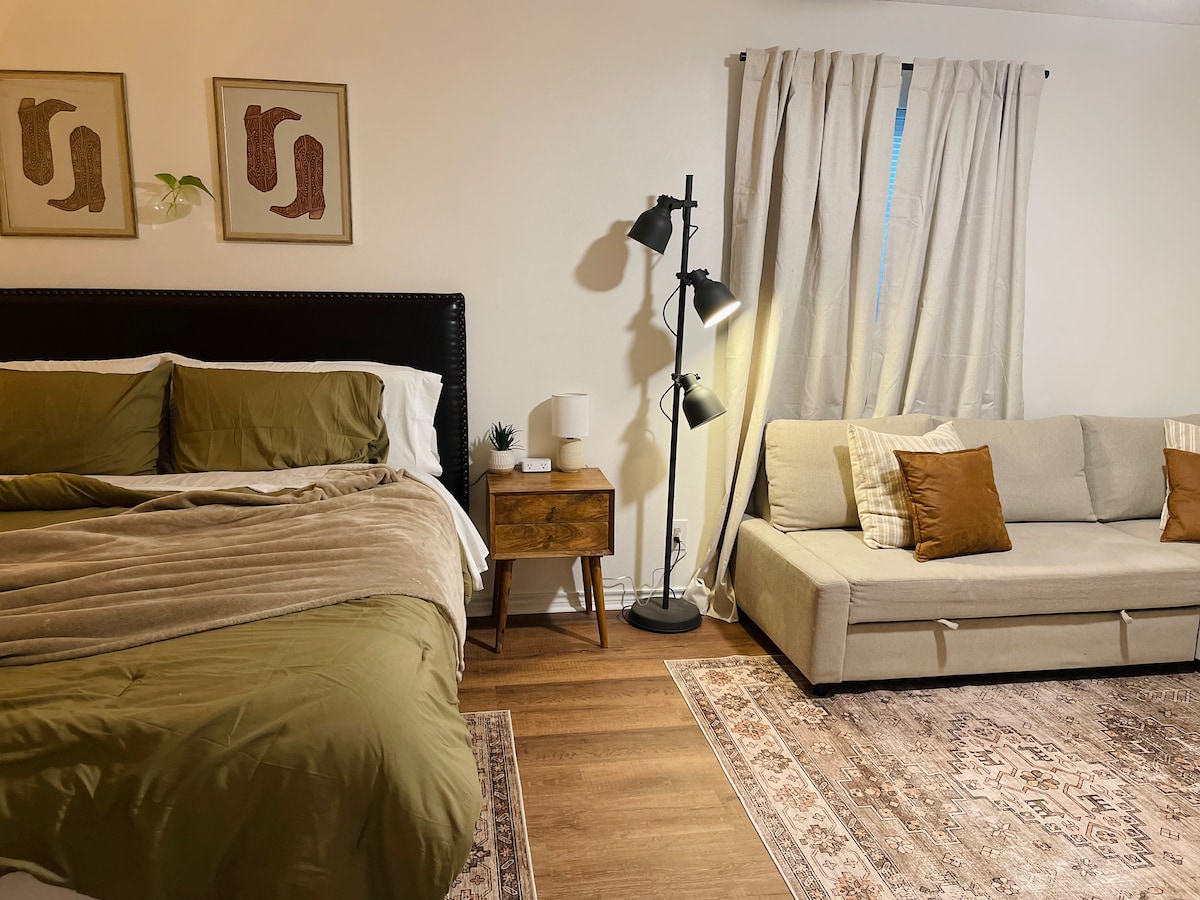 A cozy sleeping area features a spacious bed with olive green bedding and matching throw blanket. A nightstand with a small plant is adjacent, while a light gray sofa with decorative pillows sits opposite. Soft natural light filters through a window draped with light curtains.
