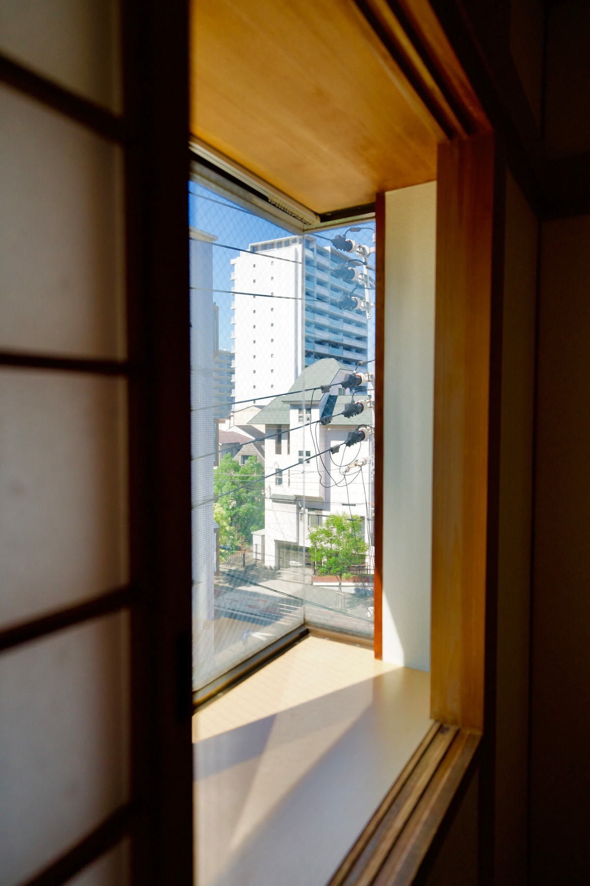 A glimpse through a wooden-framed window reveals a view of adjacent buildings and greenery outside. Sunlight illuminates the space, creating a bright and airy atmosphere. The window's design features traditional sliding panels, adding an element of cultural charm.