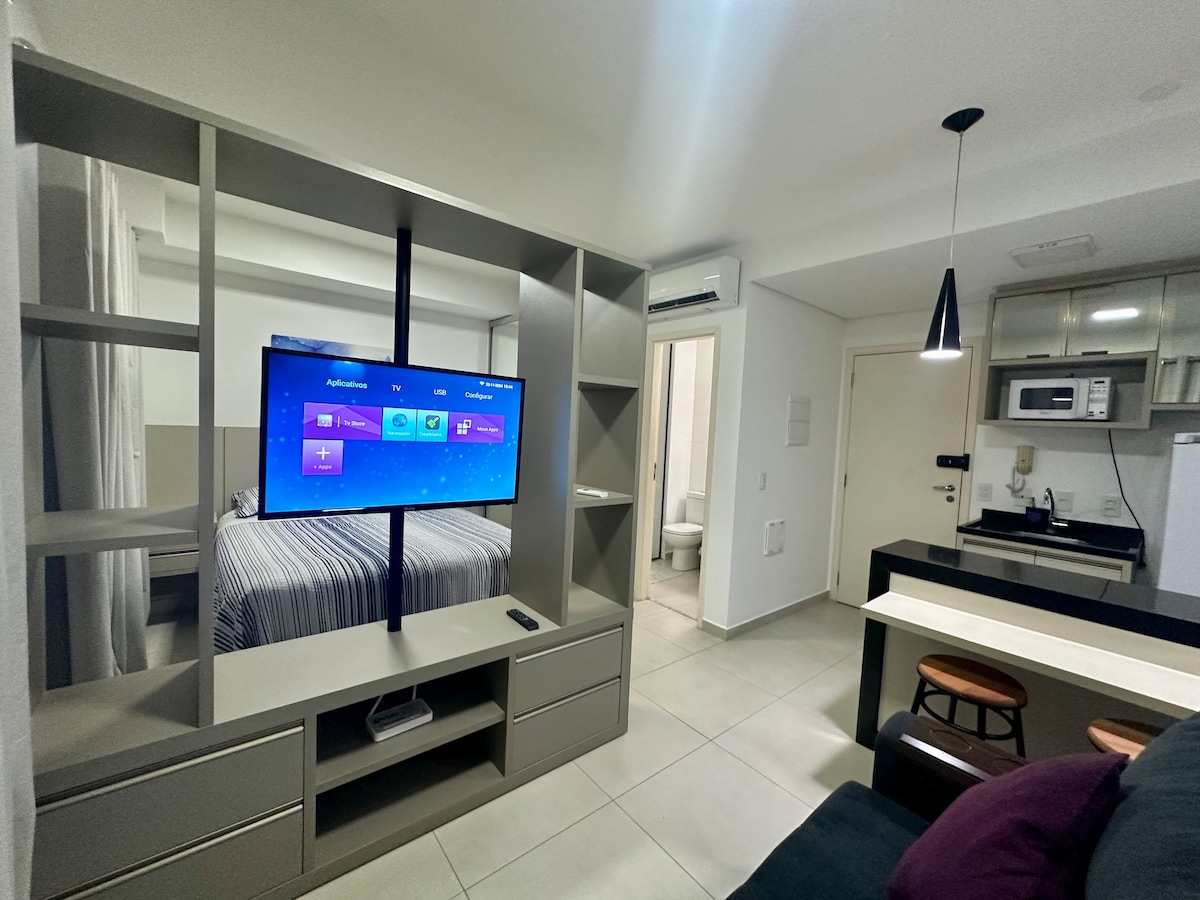 A modern living space is presented, featuring a wall-mounted TV and a sleek shelving unit. The area includes a cozy sectional sofa and a dining spot with two stools. A doorway leads to a private bathroom, while light fixtures provide general illumination.