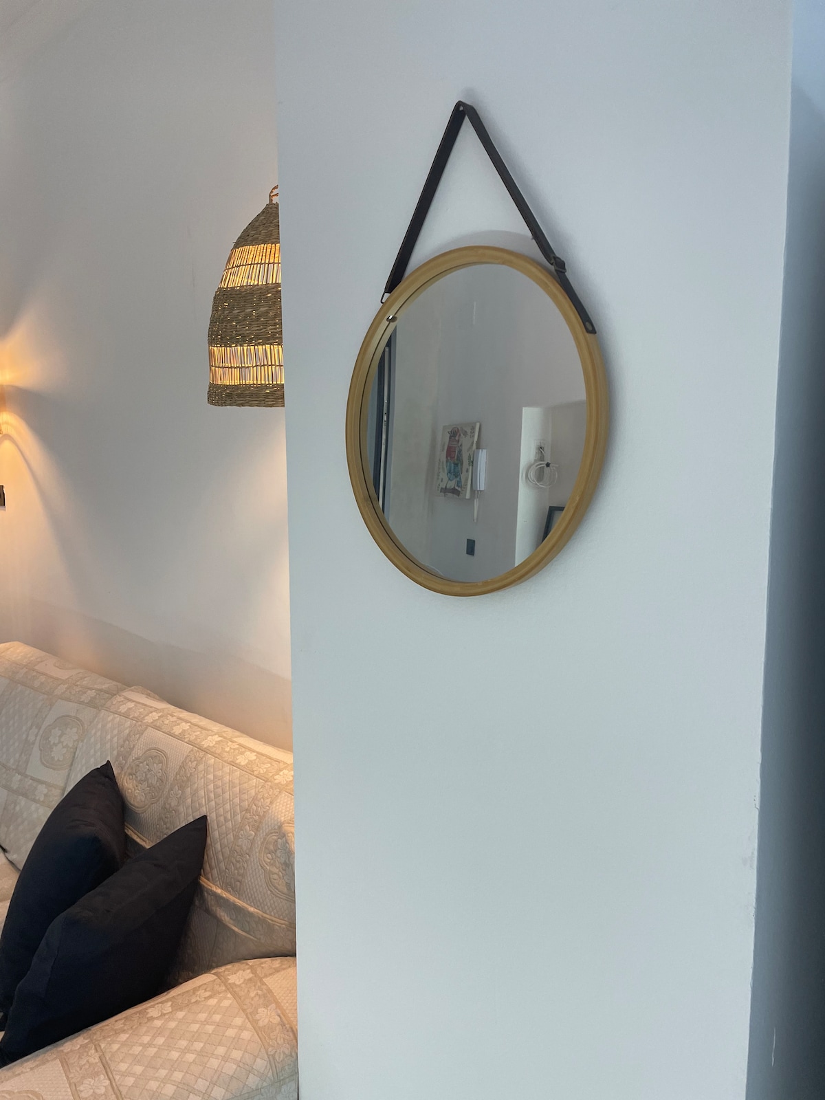 A round mirror with a gold frame is hung against a white wall. Soft lighting from a nearby lamp creates a warm ambience, while a cozy sofa with dark cushions is visible in the foreground.