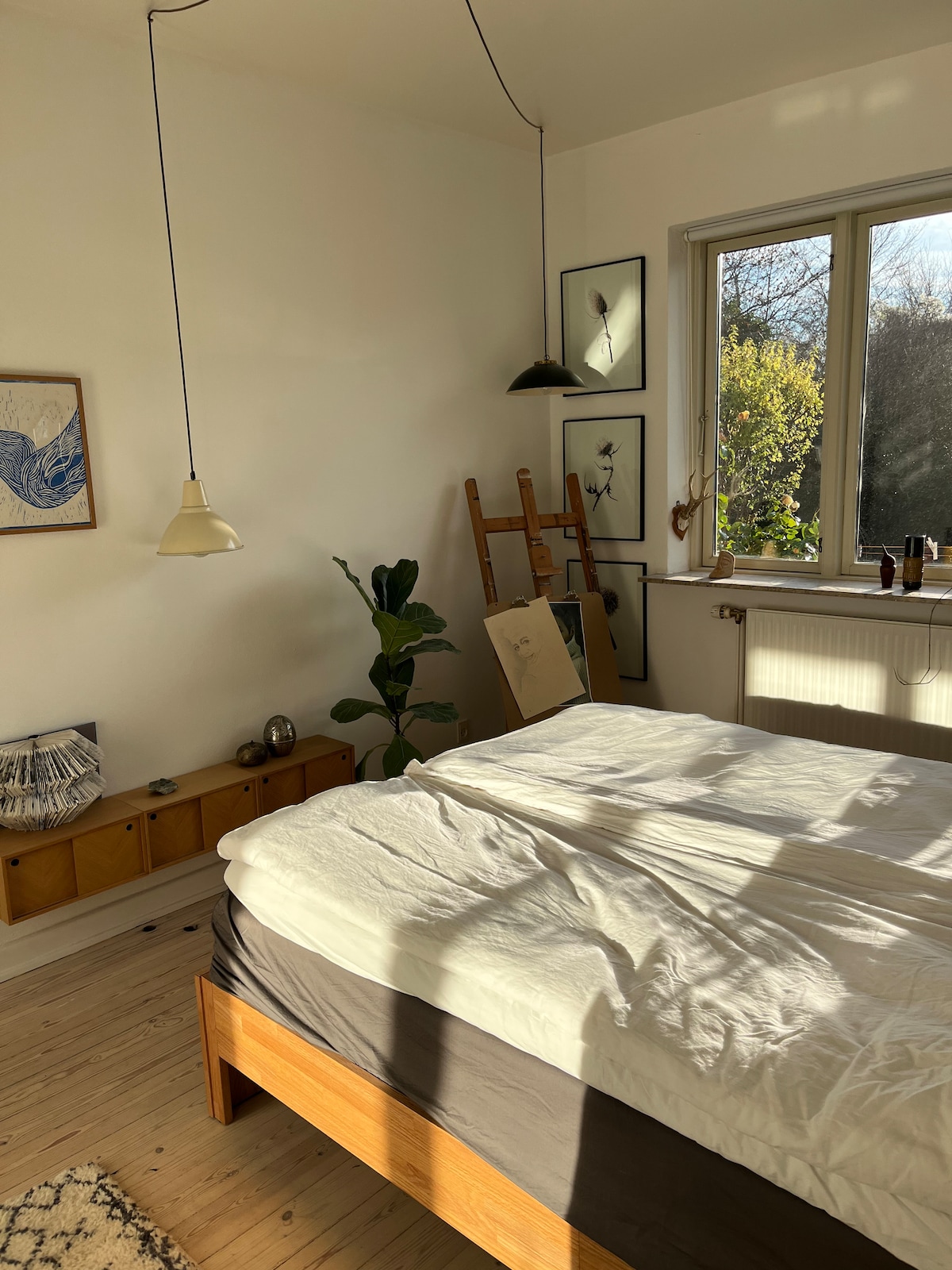 A bright bedroom features a spacious bed with a light-colored duvet. A large window allows natural light to filter in, casting soft shadows across the wooden floor. Nearby, a green plant is positioned next to a wooden shelving unit adorned with decorative items and artwork.