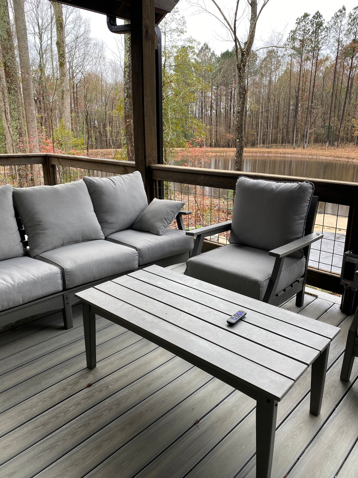A comfortable seating area is depicted on a wooden deck, featuring a gray sofa and a matching armchair. A rectangular table made of gray wood is positioned in between. The surrounding forest and a serene pond are visible in the background, enhancing the outdoor experience.