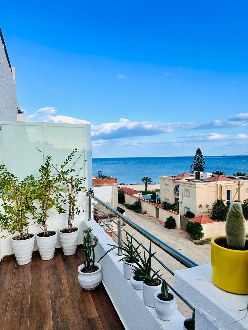 A spacious balcony is adorned with several potted plants, contributing to a vibrant outdoor space. A clear view of the sea extends beyond the railing, while the sky is brightly lit, featuring scattered clouds. Nearby buildings are visible, adding context to the serene coastal setting.