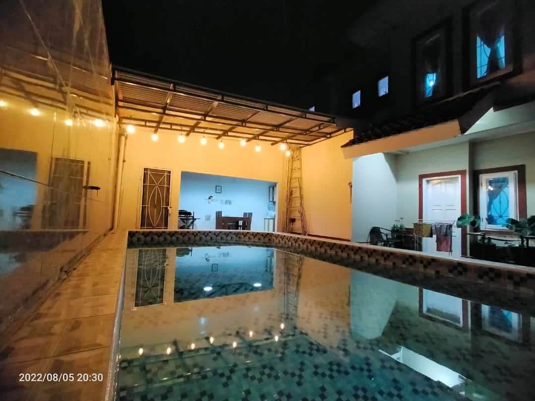 A reflective swimming pool is surrounded by a well-lit area, with soft glowing lights overhead. The indoor space features a dining table visible in the background, along with various doors leading to different rooms.