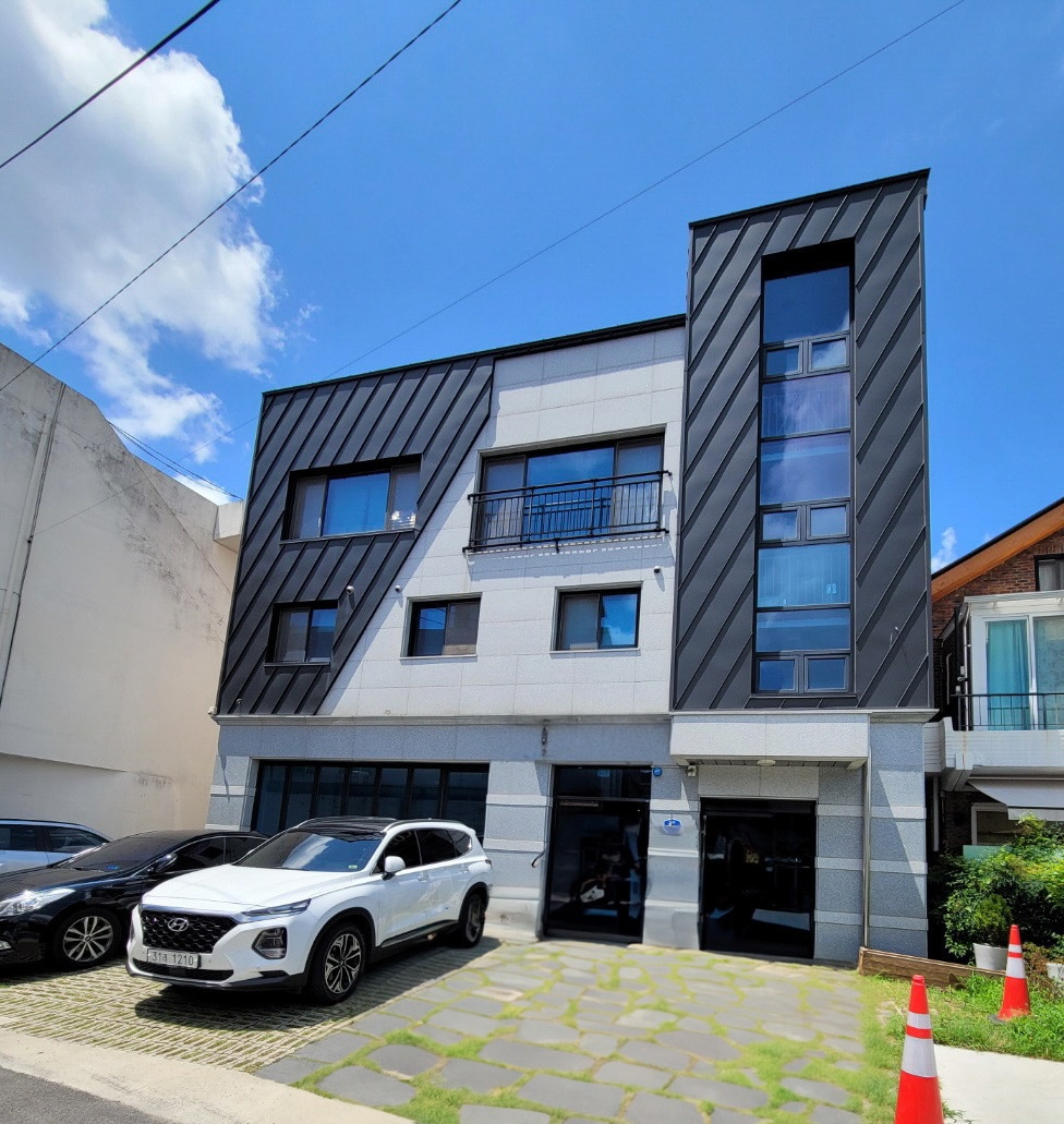 A three-story building features a modern exterior with a combination of dark and light paneling. Large windows are present, allowing natural light into the structure. Two cars are parked outside, and a landscaped stone pathway leads to the entrance.