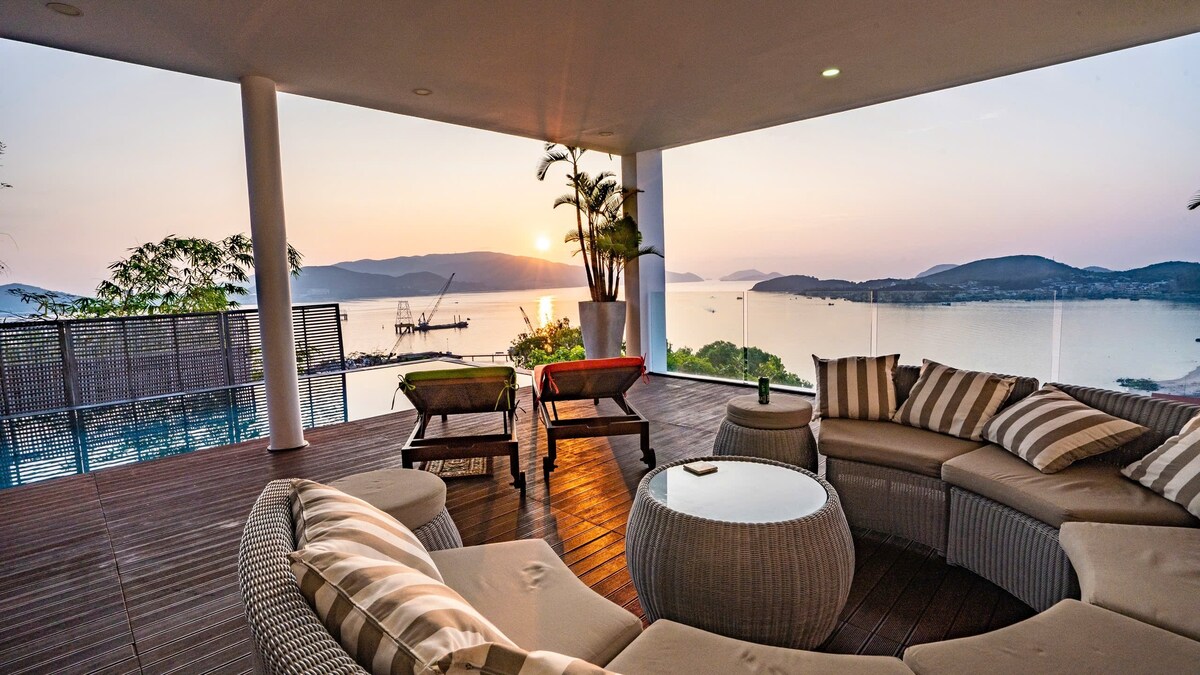 A spacious outdoor lounge area features a sectional sofa with soft cushions and a round coffee table. The scene is complemented by a stunning sunset view over the ocean, framed by lush greenery and gentle hills in the background.