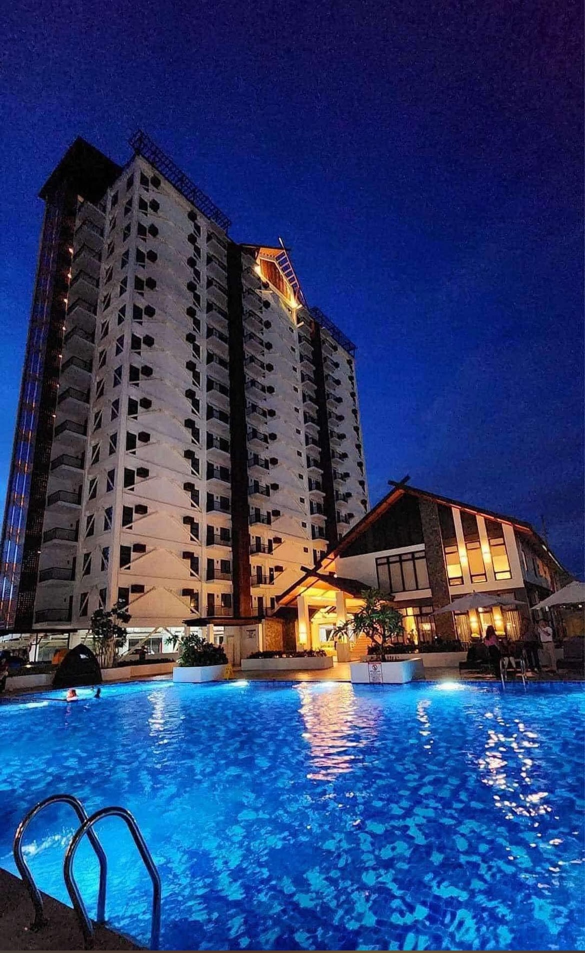 A modern condominium building is illuminated against the evening sky, showcasing a well-lit pool in the foreground. The pool reflects a calming blue hue, while the structure displays numerous balconies. A nearby poolside amenity provides an inviting atmosphere for relaxation.