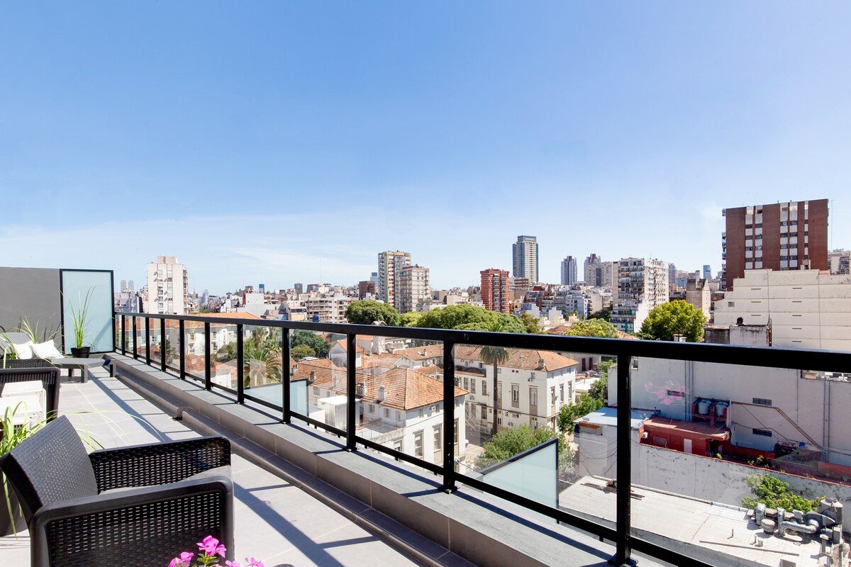 A spacious private balcony overlooks a vibrant cityscape, framed by a sleek black railing. Two lounge chairs provide a comfortable space for relaxing, while the urban horizon features a mix of buildings and greenery beneath a clear blue sky.