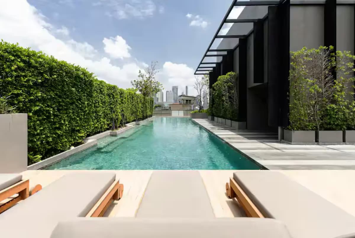 A serene rooftop sky pool stretches into the distance, framed by lush greenery and sleek modern architecture. Sun loungers are positioned along the edge, inviting relaxation while offering unobstructed views of the city skyline beyond.