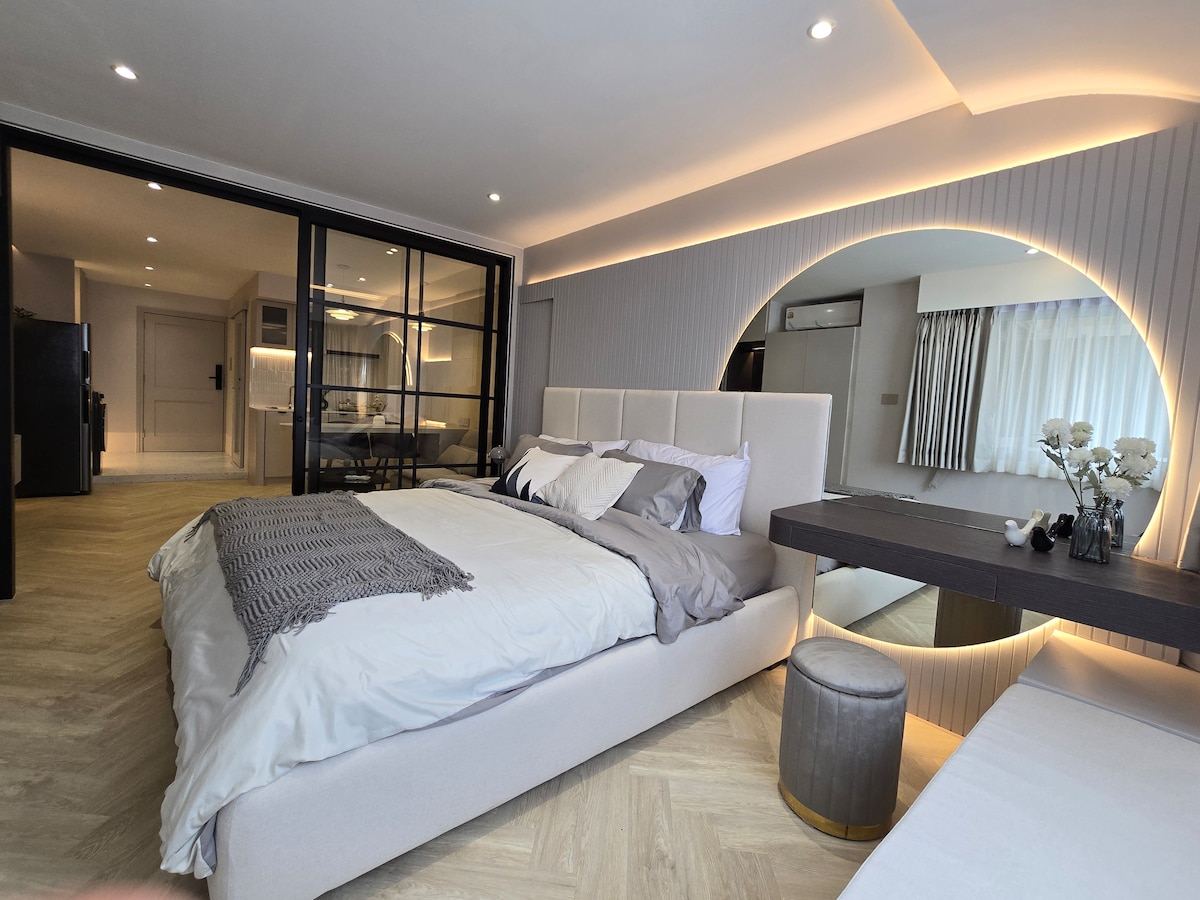 A stylish room features a soft, comfortable bed with satin bedding and decorative pillows. An inviting circular mirror reflects soft lighting. A compact seating area is positioned near a glass wall, showcasing the room's modern design and spacious layout.