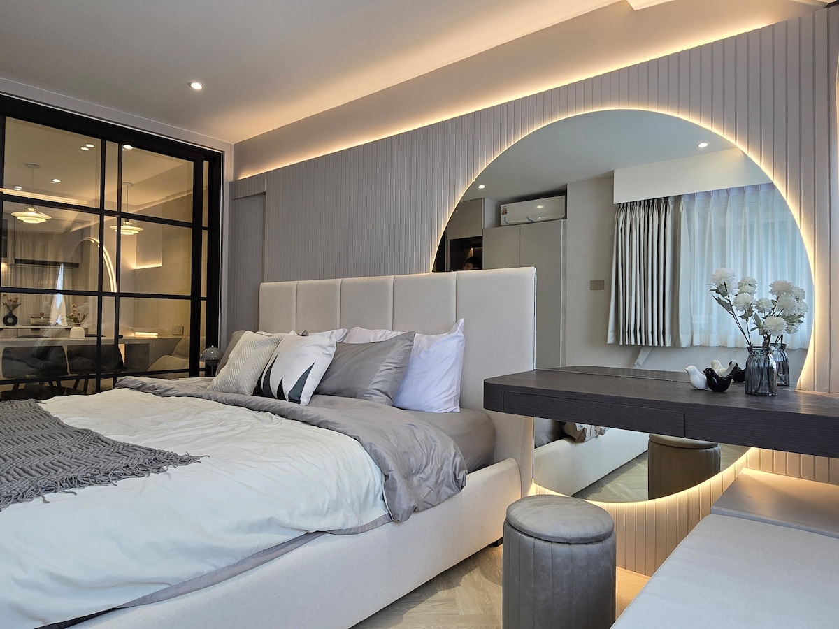 A stylish room is featured with a spacious bed dressed in soft bedding, complemented by decorative pillows. A large circular mirror is mounted on the wall, reflecting soft lighting. A modern table stands nearby, providing a functional workspace. Light filters through sheer window treatments.