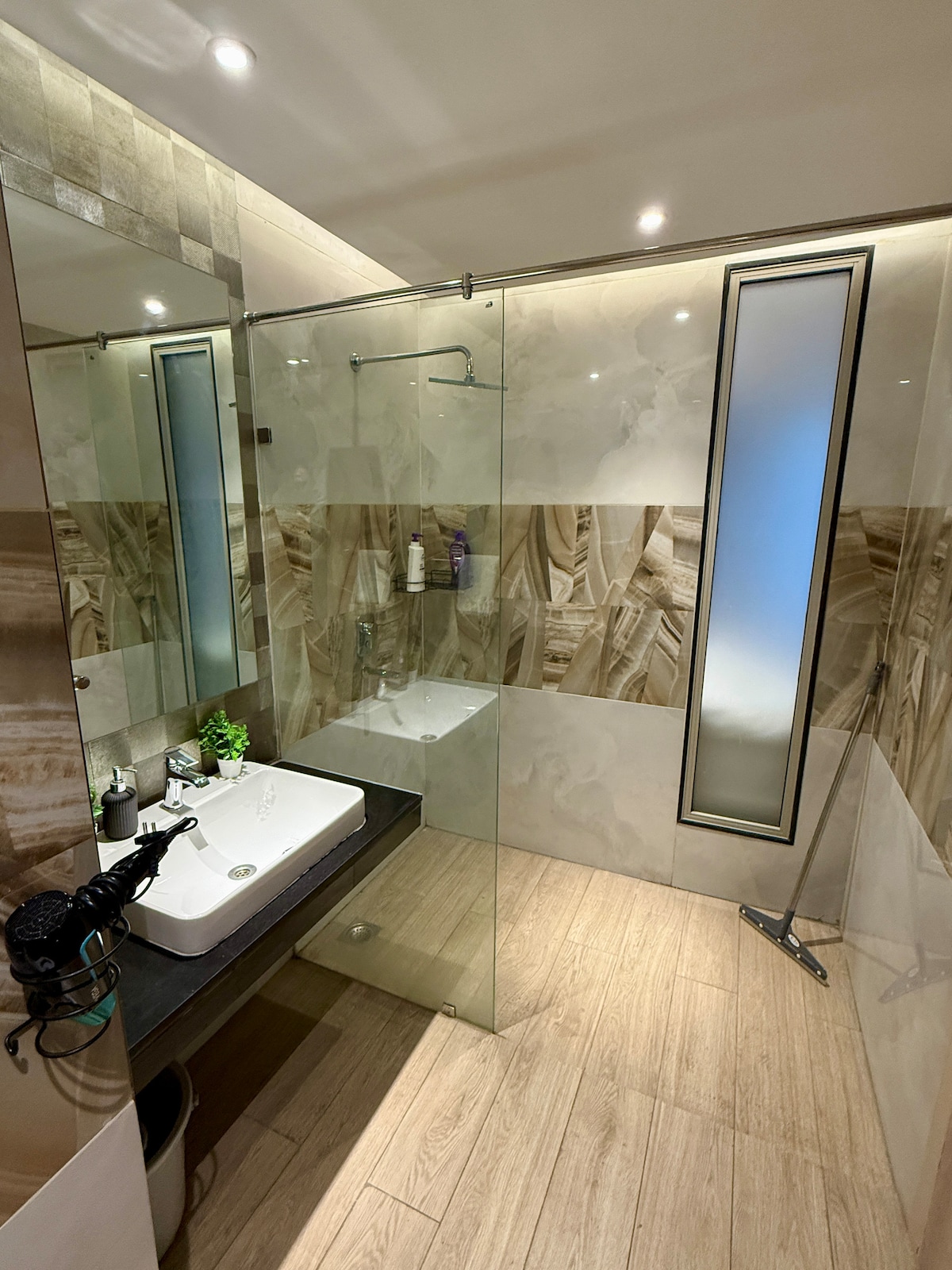 The modern bathroom features a glass-enclosed shower and a sleek sink with stylish fixtures. Neutral-toned tiles provide a clean and spacious ambiance. A large mirror enhances the area's light, while a small potted plant adds a touch of greenery.