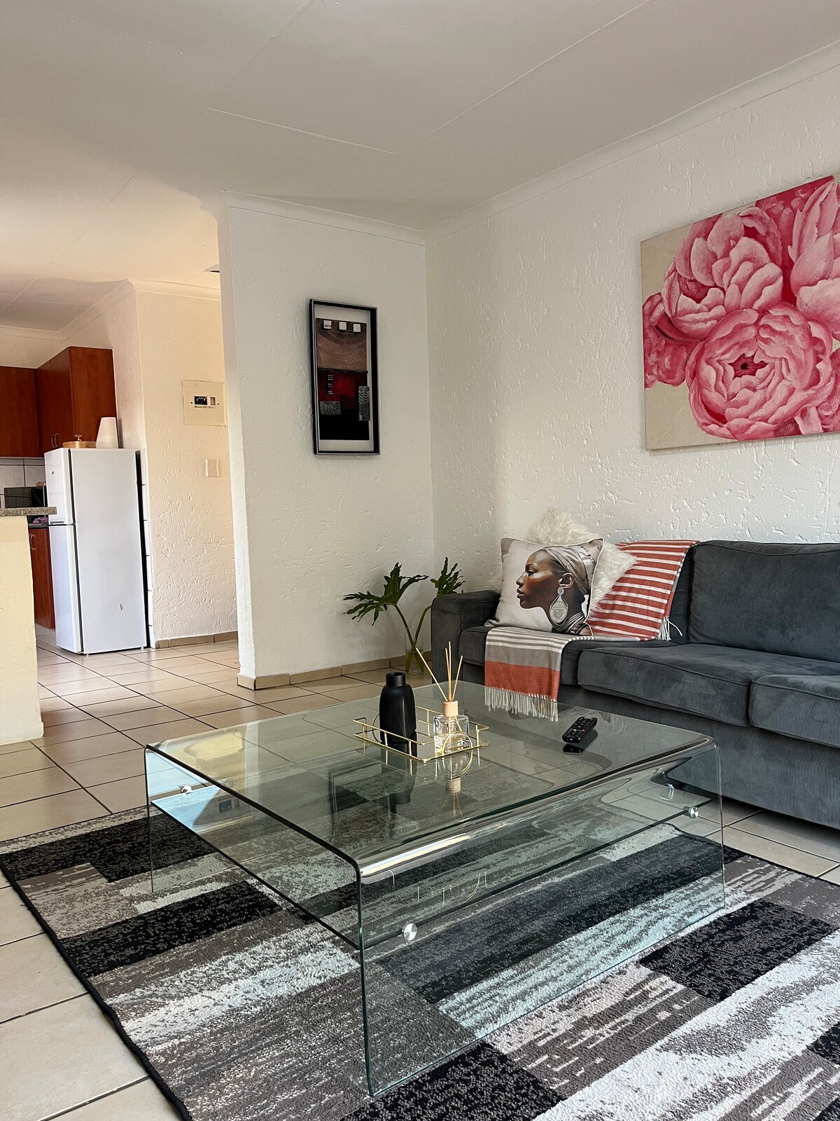 A modern living area is presented, featuring a glass coffee table atop a patterned area rug. A comfortable gray sofa is adorned with decorative pillows. A kitchen space can be seen in the background, along with a large floral art piece hanging on the wall.