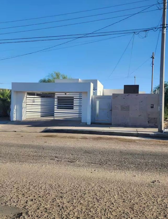 The exterior of a modern one-story home is displayed, featuring a white facade with horizontal slatted elements. A small entrance is visible on the right, and a spacious garage is positioned in the center. Clear blue skies serve as a backdrop.