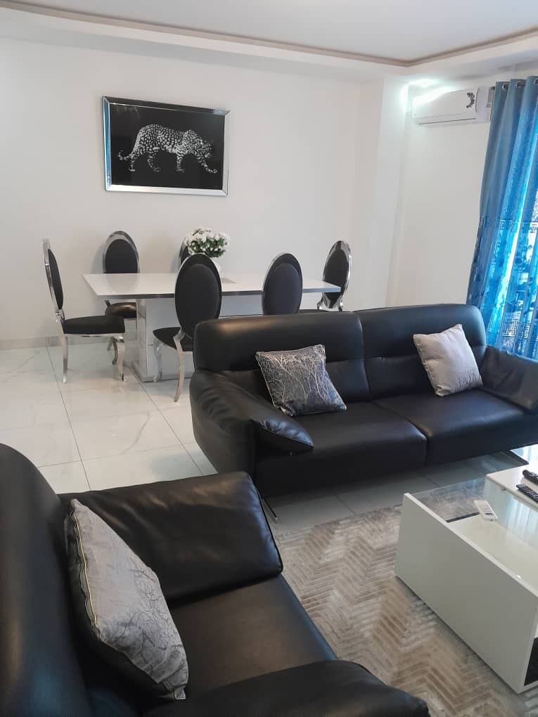 A modern living area is presented, featuring dark leather sofas with decorative pillows. A glass coffee table is situated in front of the seating. In the background, a formal dining space with a white table and six upholstered chairs is highlighted, complemented by a framed artwork on the wall.