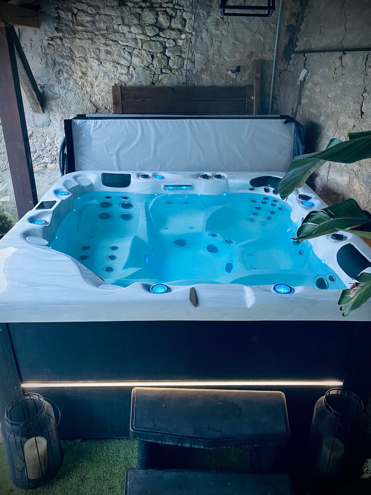 An outdoor spa is featured in a secluded area, surrounded by stone walls. The spa, with clear blue water, includes built-in seating and several jets for relaxation. Soft lighting enhances the setting, creating a serene atmosphere for unwinding.