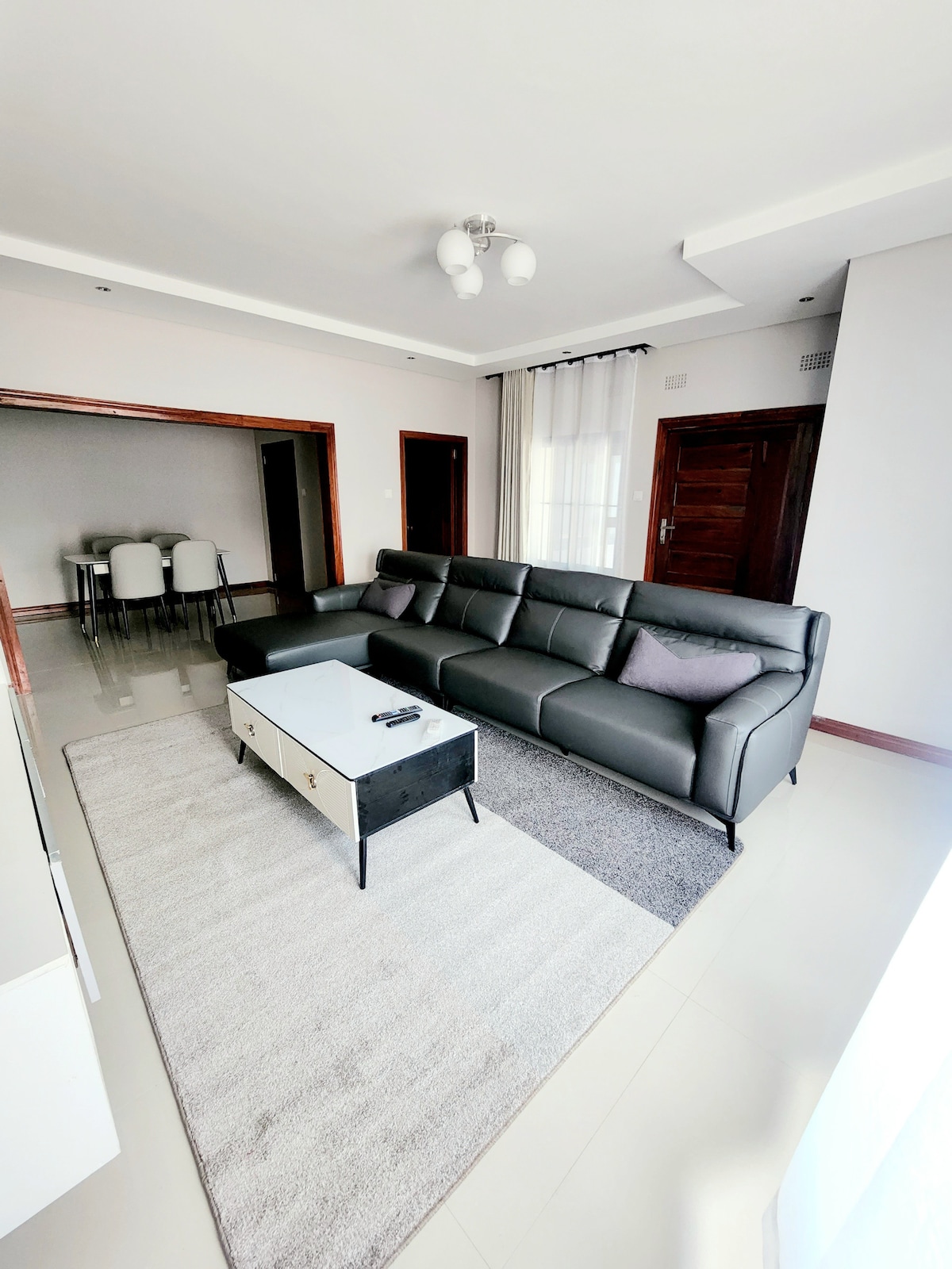 A bright living area features a dark leather sectional sofa with decorative cushions, positioned around a light-colored coffee table. A soft area rug covers the floor, while large windows offer natural light. A dining area with four chairs is visible in the background.