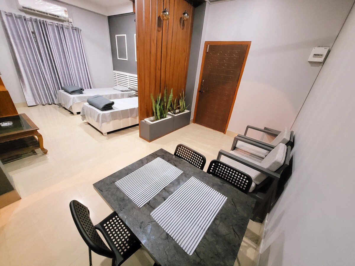 A spacious living area features a modern dining table set for four, complemented by sleek dining chairs. Two beds are neatly arranged in the background, with soft gray linens, while potted plants add a touch of greenery beside the entrance door.