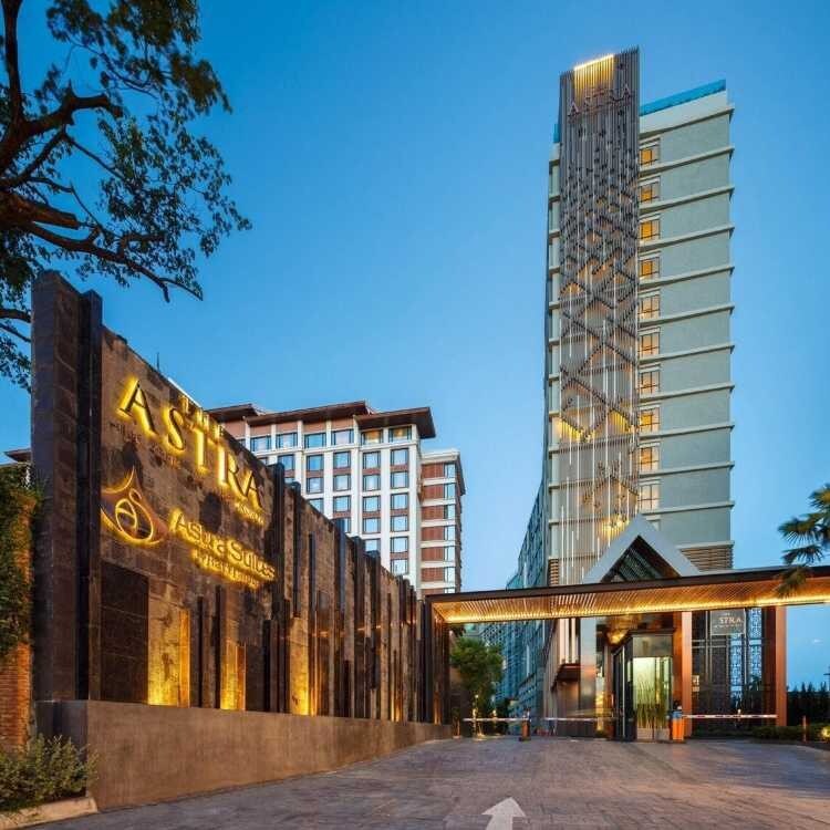 The image presents the Astra apartment building, a modern structure highlighted by a distinctive facade. The welcoming entrance is flanked by elegant landscaping, while the tall building features a unique architectural design. The sky is illuminated with soft evening light, enhancing the overall appearance.