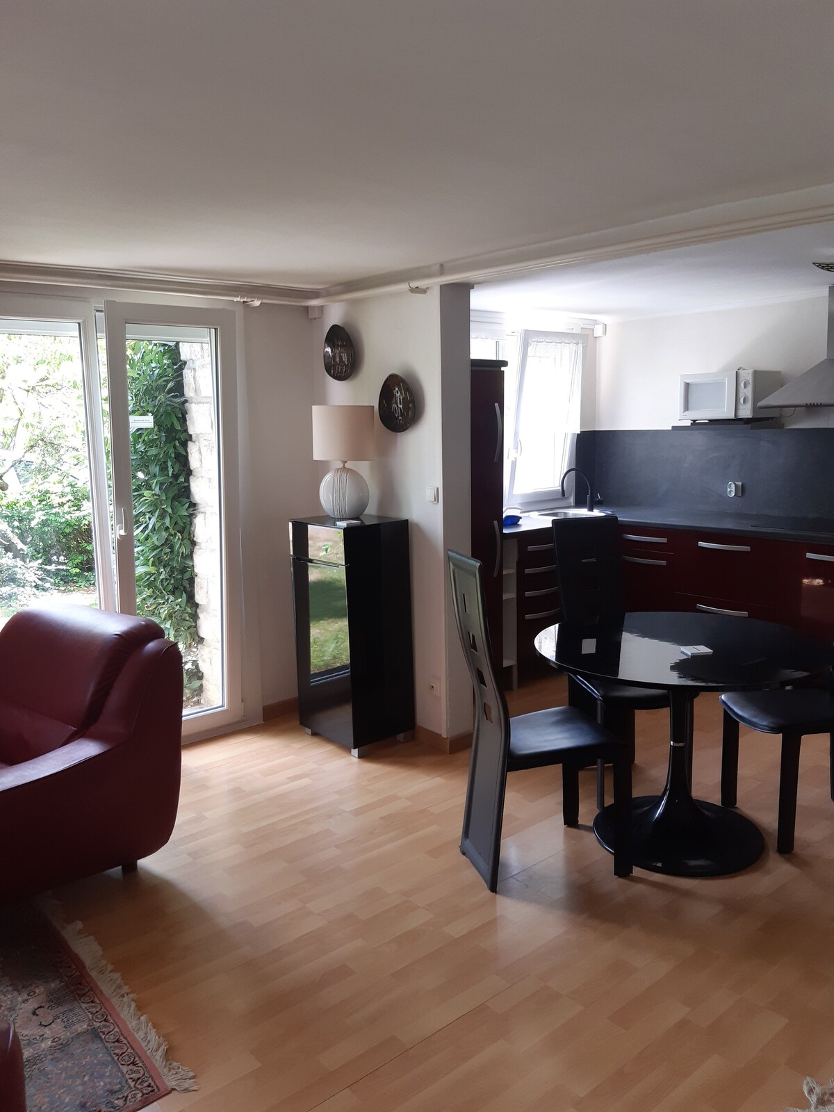 A modern living area features stylish furnishings including a red armchair and a round black dining table surrounded by four chairs. Natural light enters through large glass doors, highlighting the integrated kitchen area with dark cabinetry. A small lamp atop a black cabinet adds a soft touch.