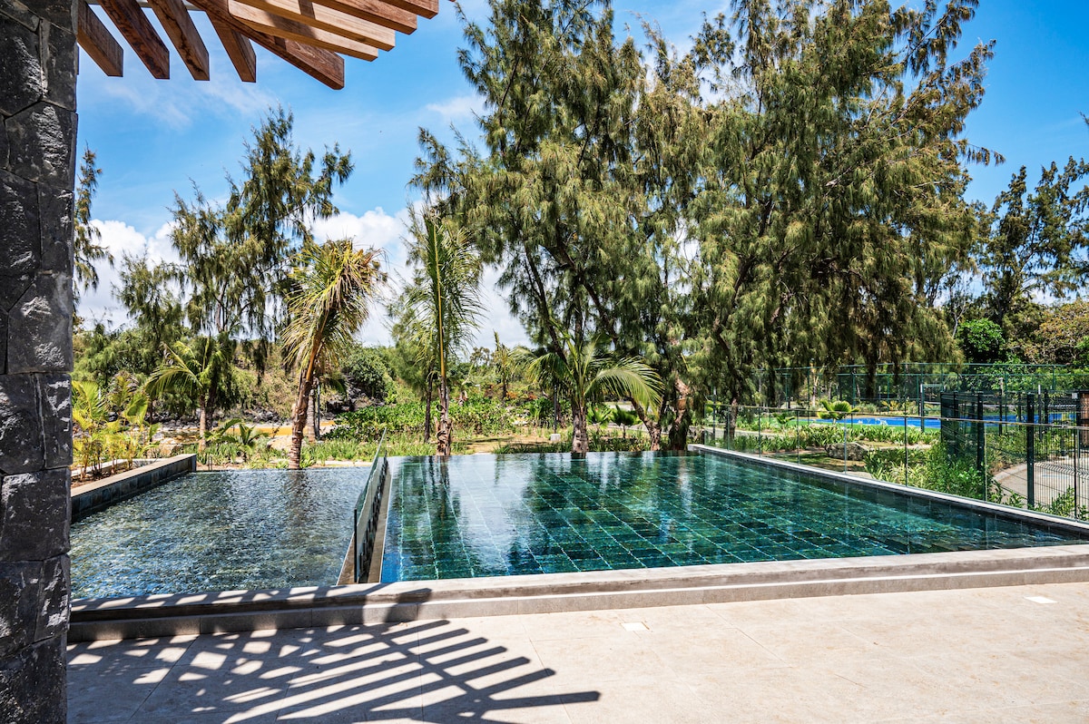 A spacious outdoor swimming pool is framed by lush greenery and palm trees, creating a serene environment. The pool's reflective surface mirrors the clear blue sky, enhancing the sense of tranquility in the area. A portion of the tennis court is visible in the background.